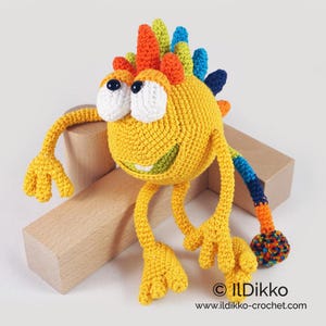 May include: A crocheted yellow monster toy with a colorful mohawk, big white eyes, and a toothy grin. The monster has long yellow arms and legs and a colorful tail.