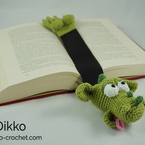 May include: Green crocheted dragon bookmark with white eyes and a pink tongue, holding a book with a red spine.
