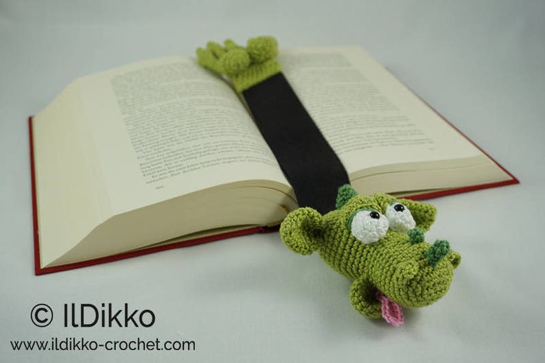 May include: Green crocheted dragon bookmark with white eyes and a pink tongue, holding a book with a red spine.