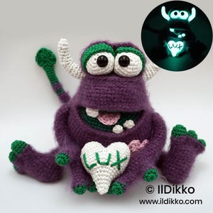 Puede incluir: Un peluche de monstruo tejido a ganchillo en color morado con cuernos blancos, ojos grandes y detalles verdes en pies y cola. Sostiene un coj&iacute;n blanco en forma de coraz&oacute;n bordado con el texto LUX. Un recuadro muestra el juguete brillando en la oscuridad.