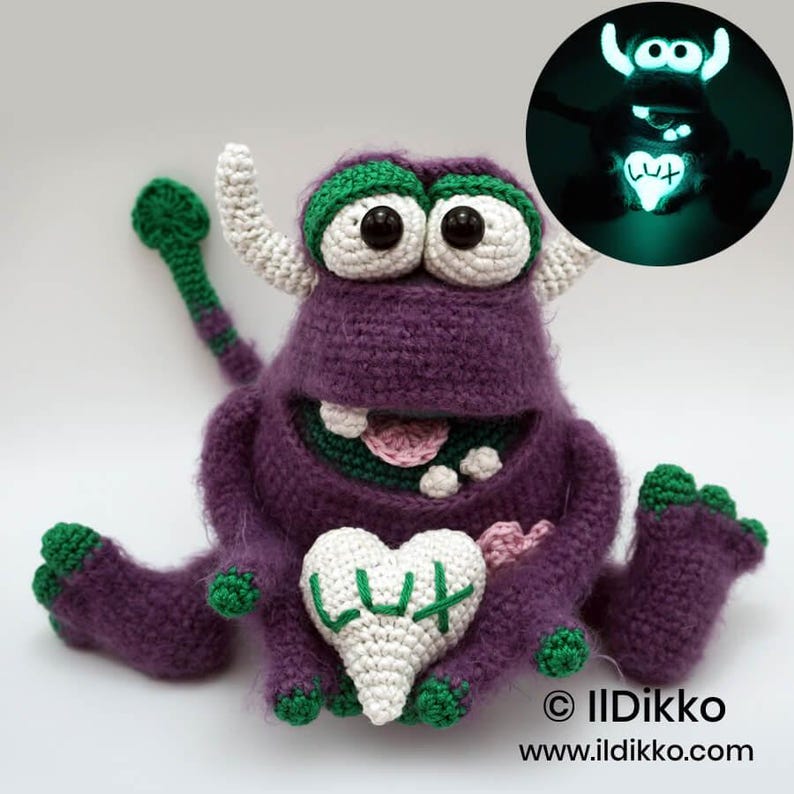 Puede incluir: Un peluche de monstruo tejido a ganchillo en color morado con cuernos blancos, ojos grandes y detalles verdes en pies y cola. Sostiene un coj&iacute;n blanco en forma de coraz&oacute;n bordado con el texto LUX. Un recuadro muestra el juguete brillando en la oscuridad.