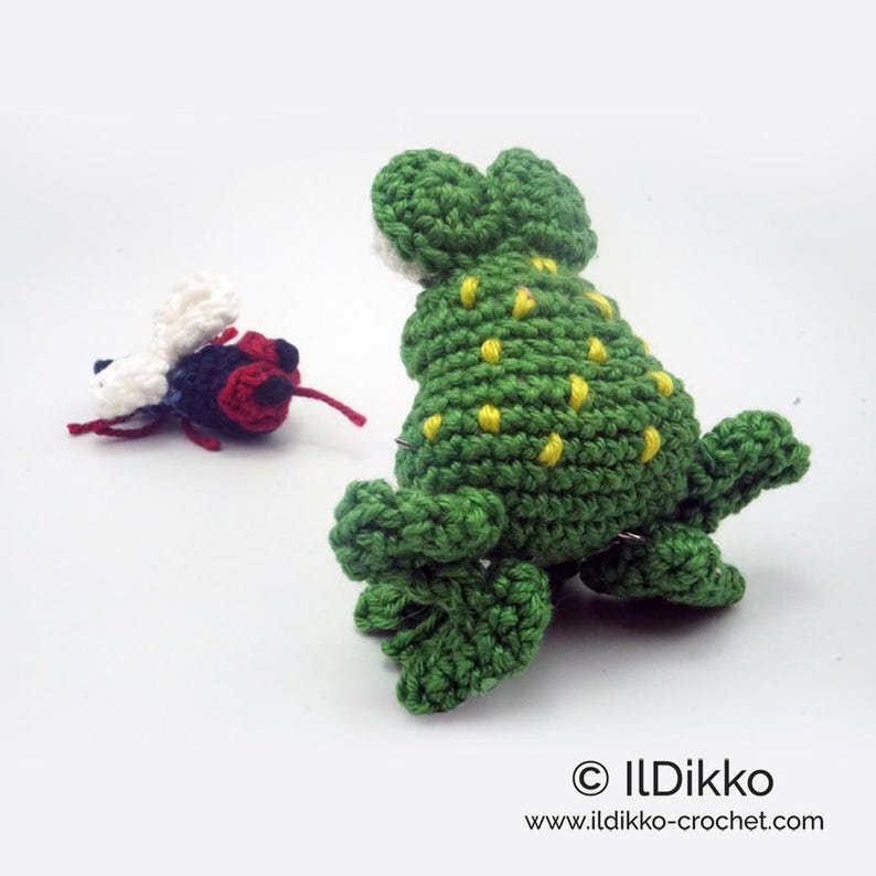 May include: A green crocheted frog with yellow spots, sitting on a white surface. A crocheted black and red fly is in the background.