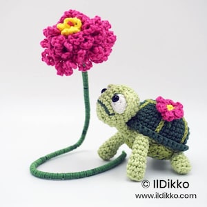 May include: A crocheted turtle with a dark green shell and light green body, featuring a small pink flower on its back. Beside it stands a tall, thin green stem topped with a large, textured pink and yellow crocheted flower. Text reads: © IlDikko www.ildikko.com