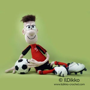 May include: A crocheted doll of a soccer player with brown hair, wearing a red jersey with the number 9 in white, black shorts, and white cleats. The doll is sitting on the ground with one hand resting on a black and white crocheted soccer ball.