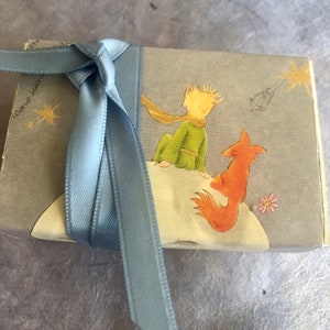 May include: A blue ribbon tied around a small book with a light blue cover. The book features an illustration of The Little Prince with a fox on a moon-like surface. The text on the cover reads "Icono Star".
