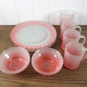 May include: A collection of frosted glass dinnerware featuring a pink ombre honeycomb pattern. The set includes a stack of round plates, two small bowls, three handled mugs, and one tall drinking glass, all with a textured, dimpled surface.