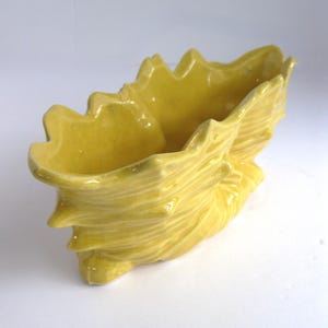 May include: A yellow ceramic seashell-shaped planter. The planter has a textured surface with ridges and a glossy finish. The interior is smooth and open, suitable for small plants or decorative items. The planter is approximately 8 inches long.