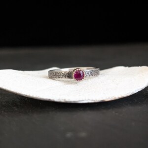 May include: A silver ring with a textured band and a round, deep red gemstone. The ring is displayed on a white, shell-shaped surface against a dark background. The gemstone is the focal point of the ring.