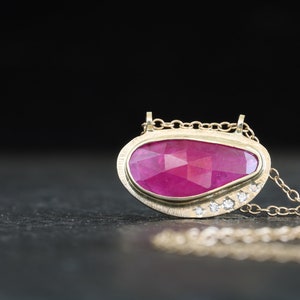 May include: A gold pendant necklace with a pink ruby gemstone set in a gold oval frame. The pendant is accented with small diamonds around the edge.