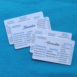 May include: Three white cards with rounded corners are stacked on a blue surface. The cards have text in a dark blue font, including the words "Margaritaville," "Islander," and "Paradise." The cards list dining, entertainment, and assistance options.