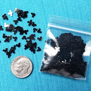 May include: A small clear plastic bag filled with black glitter shaped like Mickey Mouse heads. A small pile of the glitter is scattered on a blue surface next to a 10 pence coin for scale.