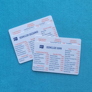 May include: Two white cards with black text listing the dining and bar options on the Norwegian Breakaway and Norwegian Dawn cruise ships. The cards are on a blue surface.