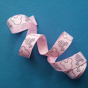 Op de afbeelding: Roze lint met een herhaald patroon van Mickey Mouse-silhouetten en tekst. Het lint is gekruld en ligt op een turquoise blauw oppervlak. Geschikt voor knutselen, cadeauverpakking of decoratie.