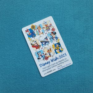 May include: A white Disney Wish 2025 card featuring Disney characters in sailor outfits. The card has the text "Pull Light Card up slightly to disengage before leaving room". The card is on a blue surface.