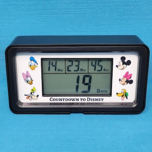 May include: Black digital countdown clock with a white face. The clock displays "14 Hours 23 Minutes 45 Seconds 19 Days" and features cartoon images of Mickey Mouse, Minnie Mouse, Donald Duck, Daisy Duck, and Goofy. The clock reads "Countdown to Disney".