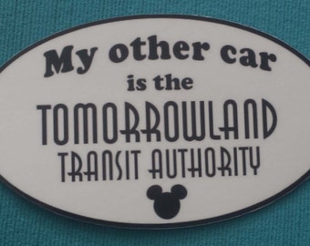 Könnte beinhalten: Weißer ovaler Aufkleber mit schwarzem Text, der "My other car is the TOMORROWLAND TRANSIT AUTHORITY" lautet, mit einer schwarzen Mickey-Maus-Silhouette unten.
