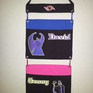 May include: A black fabric wall organizer with three pockets. The top pocket has a red and white Disney cruise line logo. The middle pocket has a blue top border and the name "David" printed on it. The bottom pocket has a pink top border and the name "Jenny" printed on it.