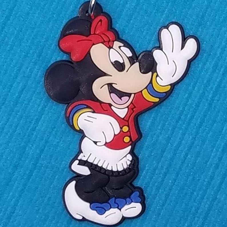 May include: A red and black rubber figurine of Mickey Mouse's girlfriend, Minnie Mouse, wearing a red jacket with yellow buttons and a white skirt. She is waving with her right hand and has a red bow in her hair.