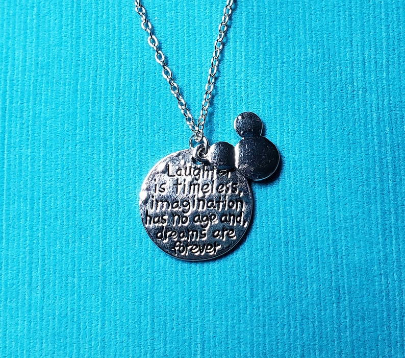 May include: Silver-toned necklace with a pendant featuring the quote "Laughter is timeless, imagination has no age and dreams are forever." Includes a small Mickey Mouse head charm. The chain is delicate, and the background is a solid teal color.