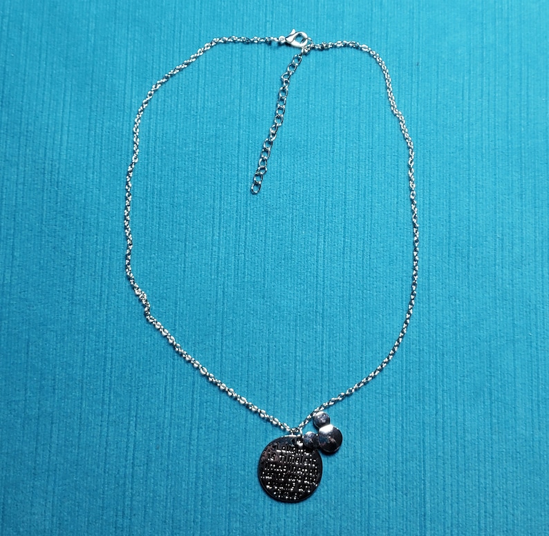 May include: Silver-toned necklace with a delicate chain and a round pendant. The pendant has etched text and is accompanied by a smaller, heart-shaped charm. The necklace is displayed against a turquoise background.