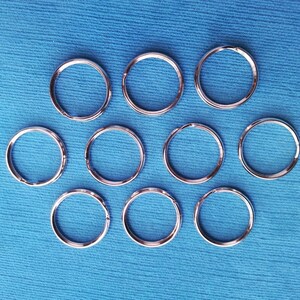 May include: A collection of ten silver-colored key rings arranged on a blue fabric surface. The key rings are circular and made of metal. The background is a solid, light blue color.