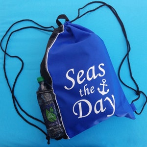 May include: A blue drawstring backpack with the phrase "Seas the Day" in white script and an anchor graphic. The bag has a black and white trim and a mesh side pocket holding a water bottle. Black drawstring cords.