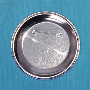 May include: The back of a silver metal button with a pin attached.