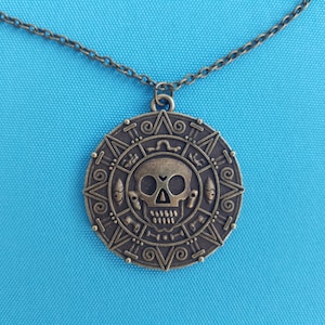 May include: A gold-toned necklace with a round pendant featuring a skull and crossbones design. The pendant has intricate details and a dark brown patina.
