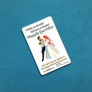 May include: A white card with a cartoon image of Ariel and Prince Eric from the Disney movie The Little Mermaid. The text on the card reads "While we devotin' full time to floatin' Happily Ever After". The card also includes instructions to "Pull Light Card up slightly to disengage before leaving room" and a note that it was "Created by PUCE DUETED Stitches".