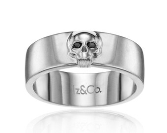 May include: A silver ring featuring a skull design with black accents on the eyes. The ring has a wide band and the text "lz&Co." is engraved on the inside. The ring is displayed on a white background.