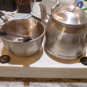 May include: A white plastic toy stove with two black circular knobs, featuring a small silver metal saucepan with a black handle and a silver metal tea kettle with a black handle and a lid, arranged on the stovetop surface for play.