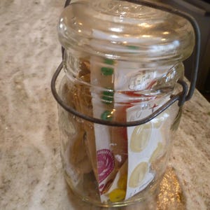 May include: A clear glass jar with a wire handle and lid, filled with packets. The jar sits on a speckled countertop. The packets have colorful designs and text, including images of lemons.