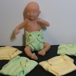 May include: A realistic baby doll wearing a green cloth diaper secured with safety pins. Four additional cloth diapers are shown in the image, two green and two yellow.