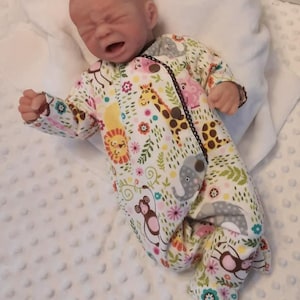 May include: A realistic baby doll with a distressed expression, wearing a white and multi-colored sleeper with a jungle animal print. The sleeper has a black trim and a zipper closure.