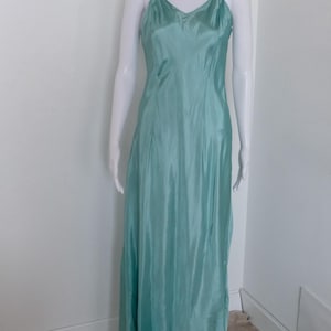 May include: A floor-length, sleeveless slip dress in a solid seafoam green color. The garment features a V-neckline, thin spaghetti straps, and a smooth, satin-like finish that drapes down to the floor. The dress is displayed on a white mannequin.