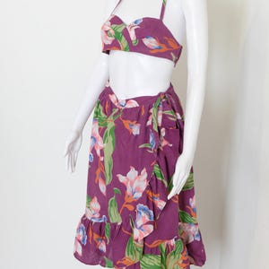 May include: A two-piece purple floral print outfit. The halter top and wrap skirt feature a vibrant pattern of white, pink, orange, and green flowers and leaves. The skirt has a ruffled hemline, adding a touch of flair to the ensemble.