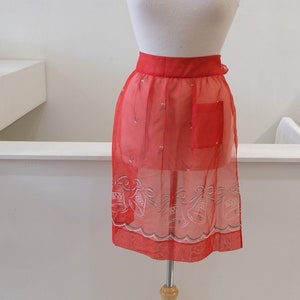 May include: A red sheer apron with a white bell pattern and a single pocket. The apron is displayed on a wooden mannequin.