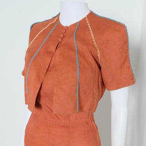 May include: A rust-colored dress with a matching short-sleeved jacket. The jacket has a unique design with a draped front and decorative stitching in light blue and yellow. The dress appears to be made of a lightweight fabric.