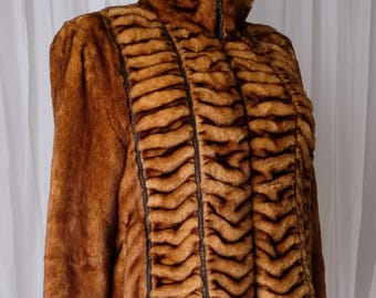 May include: A brown faux fur jacket with a high collar and a zipper closure. The jacket features vertical panels with a textured, striped pattern in shades of brown and black. The sleeves are long and the jacket is displayed on a mannequin.