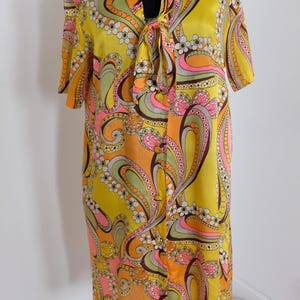 May include: A vintage-style dress in a vibrant yellow hue, adorned with a paisley pattern in shades of orange, pink, green, and brown. The dress features short sleeves, a tie-neck detail, and a knee-length cut, showcasing a retro aesthetic.