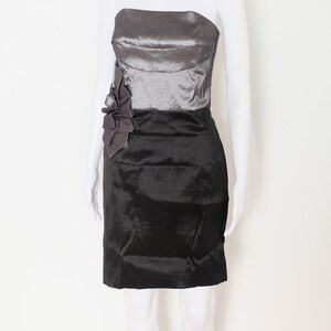 May include: A strapless, two-tone dress on a mannequin. The bodice is a shimmering silver-gray, while the skirt is a solid black. A dark gray fabric flower detail adorns the left side of the bodice. The dress is knee-length.