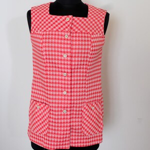 May include: A vintage red and white gingham vest with a square neckline and two front pockets. The sleeveless top has a button-up front with white buttons. The vest is displayed on a black mannequin.