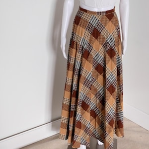May include: A long, patchwork skirt in shades of brown, tan, and cream, displayed on a mannequin. The skirt features a variety of plaid and geometric patterns. The mannequin also wears a white, fitted top. The skirt is a maxi length.
