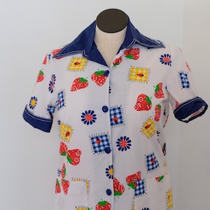 May include: A white shirt with a colourful strawberry and patchwork design. The shirt has a navy blue collar and cuffs. The pattern includes red strawberries, blue flowers, and yellow gingham squares, giving a vintage, whimsical look.