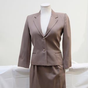 May include: A brown skirt suit displayed on a mannequin. The suit includes a blazer with two buttons and a skirt. The blazer has two front pockets. The suit is a solid color and appears to be made of a smooth fabric. The mannequin is on a metal stand.