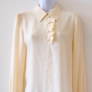 May include: A cream-colored, long-sleeved blouse with a ruffled collar and a button-up front. The blouse has a subtle polka dot pattern.