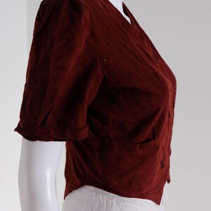 K&ouml;nnte beinhalten: Eine weinrote Bluse mit kurzen &Auml;rmeln, V-Ausschnitt und Knopfverschluss. Die Bluse hat zwei kleine Taschen und wird auf einer wei&szlig;en Schaufensterpuppe pr&auml;sentiert. Die &Auml;rmel sind umgeschlagen, und der Stoff scheint strukturiert zu sein.