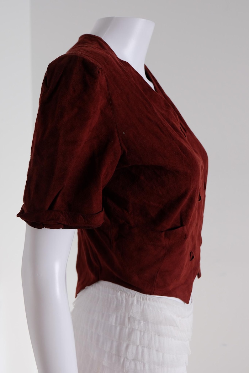 K&ouml;nnte beinhalten: Eine weinrote Bluse mit kurzen &Auml;rmeln, V-Ausschnitt und Knopfverschluss. Die Bluse hat zwei kleine Taschen und wird auf einer wei&szlig;en Schaufensterpuppe pr&auml;sentiert. Die &Auml;rmel sind umgeschlagen, und der Stoff scheint strukturiert zu sein.