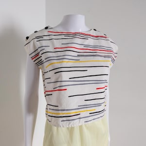 May include: A white top with a horizontal stripe pattern in red, black, gray, and yellow. The top has short sleeves and small black buttons on the shoulders. The bottom portion is a light yellow color.
