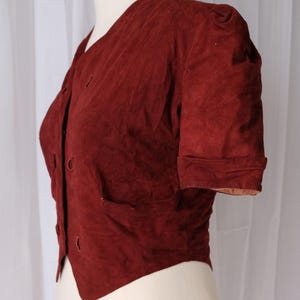 K&ouml;nnte beinhalten: Burgunderfarbene Wildlederjacke mit kurzen &Auml;rmeln und Knopfleiste. Die Jacke hat einen V-Ausschnitt und eine kleine Tasche auf der linken Seite. Die &Auml;rmel sind umgeschlagen und die Jacke wird auf einer Schaufensterpuppe pr&auml;sentiert.